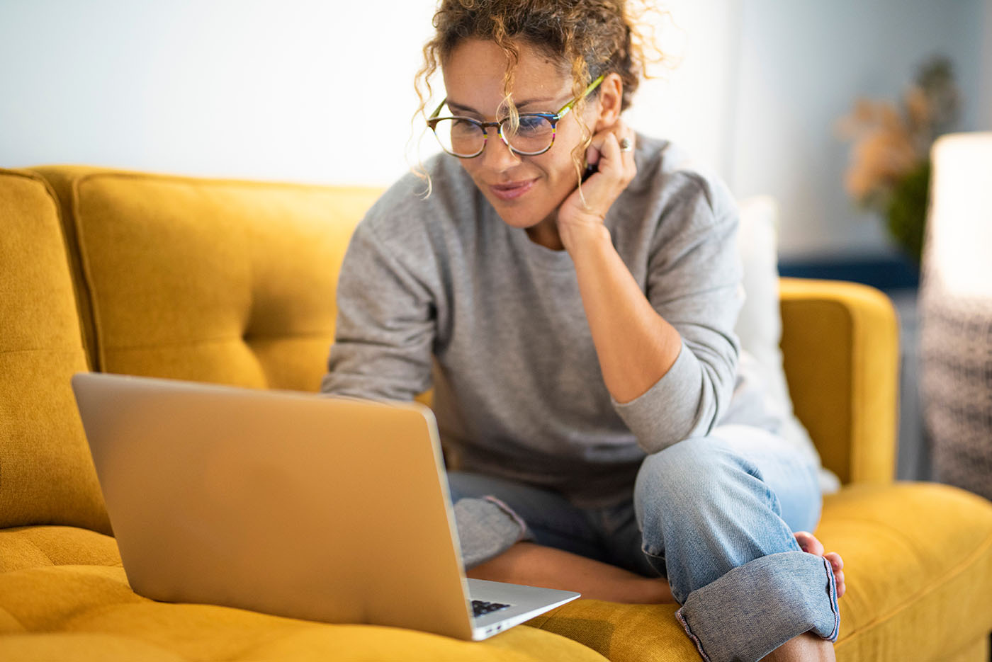 Frau sitzt auf gelbem Sofa und arbeitet mit einem Laptop.