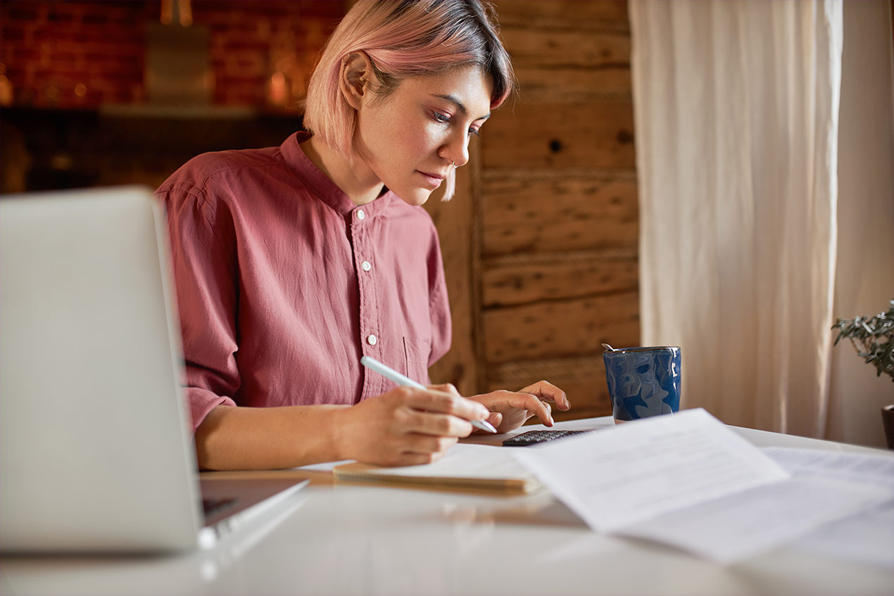 Eine Frau sitzt am Schreibtisch und macht sich Notizen. Neben ihr auf dem Schreibtisch steht ein Computer.