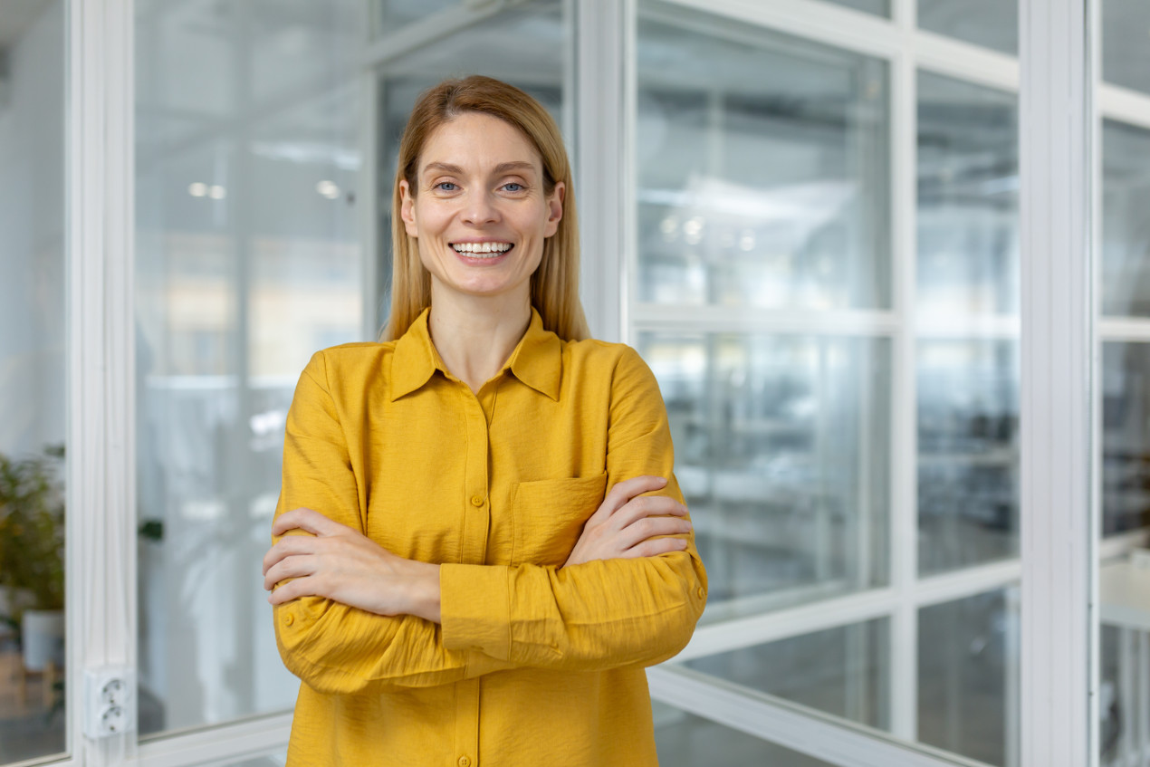 Blonde Frau mit gelber Bluse im Büro lächelt Sie an