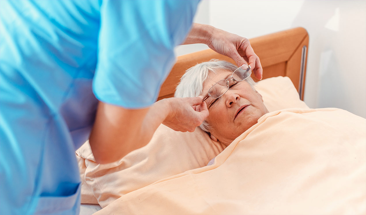 Eine Pflegerin nimmt einer liegenden Seniorin die Brille vom Gesicht