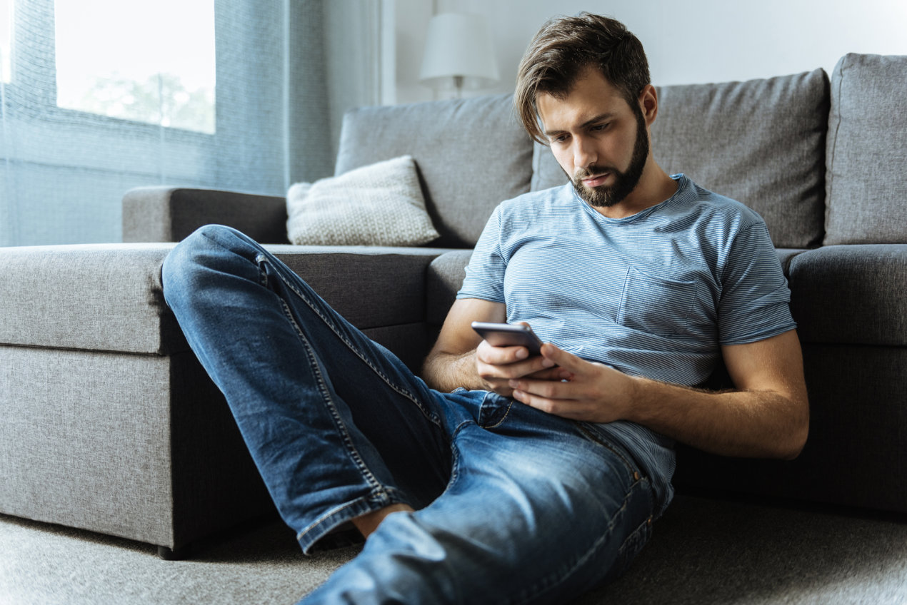 Mann mit gedrücktem Gesichtsausdruck sitzt vor dem Sofa und schaut in sein Smartphone