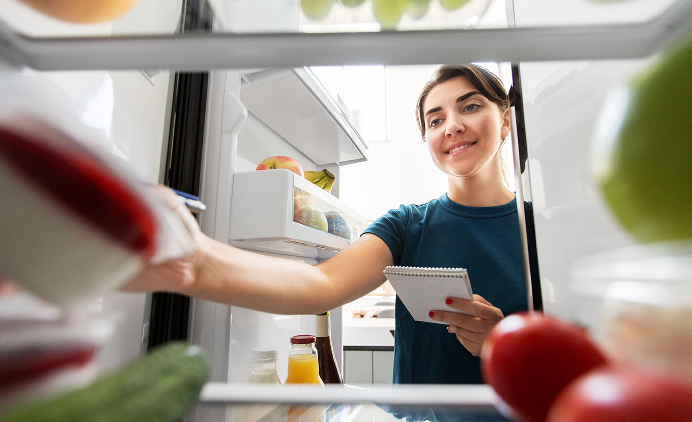 Eine Frau die mit einem kleinen Block in der Hand in den Kühlschrank schaut