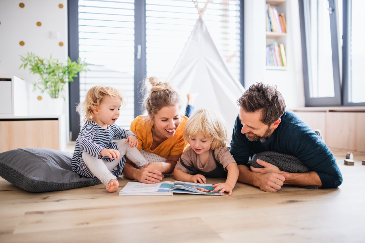 Familie mit zwei kleinen Kindern liegt auf dem Boden und schaut sich ein Heft an.